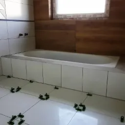 Białe płytki podłogowe w łazience, w trakcie układania, z użyciem zielonych klinów poziomujących, widoczna wanna obudowana białymi panelami oraz fragment ściany z drewnianą okładziną i oknem.