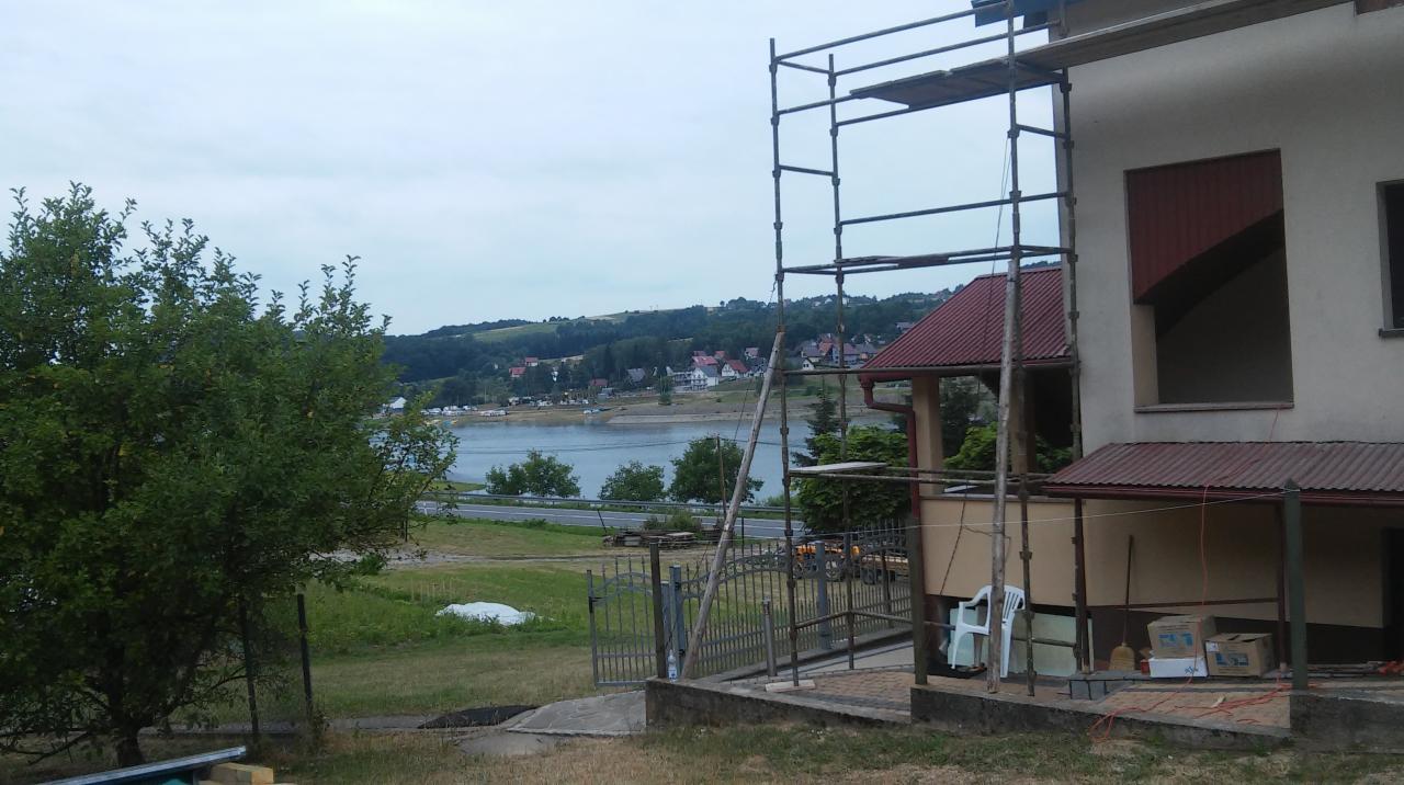 Dom w trakcie prac remontowych z widocznym rusztowaniem, z oknami bez wykończenia i widokiem na jezioro w tle.