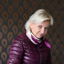 Uśmiechnięta kobieta w fioletowej kurtce na tle ściany w brązowe wzory. Portret w ujęciu od dołu, z delikatnym uśmiechem i jasnymi włosami.