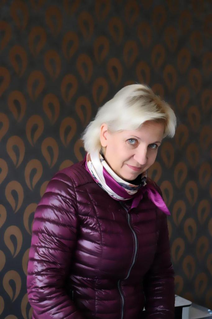 Uśmiechnięta kobieta w fioletowej kurtce na tle ściany w brązowe wzory. Portret w ujęciu od dołu, z delikatnym uśmiechem i jasnymi włosami.