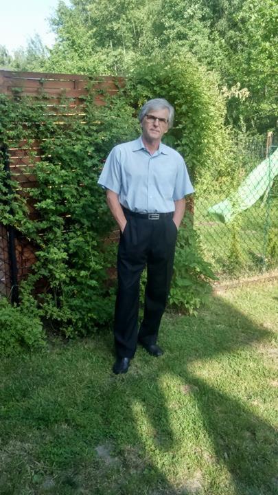 Mężczyzna w okularach, jasnej koszuli i ciemnych spodniach stoi na trawniku przed drewnianym płotem porośniętym zielenią.