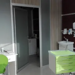 Wnętrze gabinetu stomatologicznego z mikroskopem zabiegowym, zielonym fotelem i biurkiem z przyborami biurowymi.