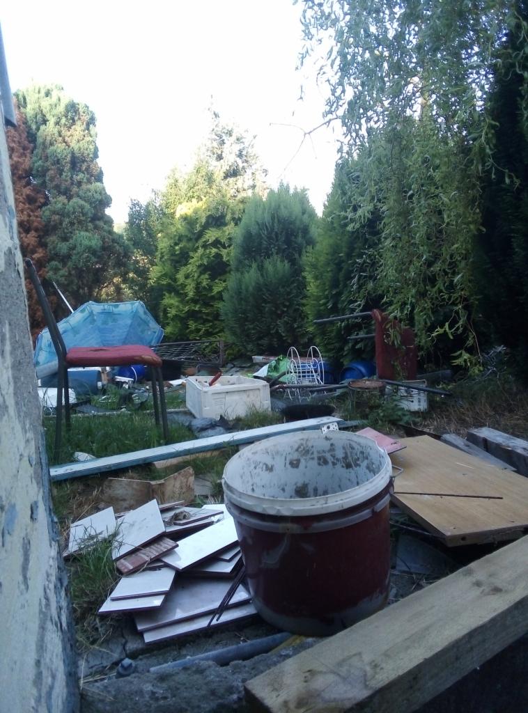 Zaniedbany ogród z porozrzucanymi materiałami budowlanymi, wiadrem po farbie, płytkami i starymi meblami na tle gęstych drzew i krzewów.