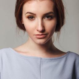 Veludo - Portret młodej kobiety z rudymi włosami upiętymi do góry, z delikatnym makijażem oczu w odcieniach złota i brązu, ubrana w bluzkę w prążki o pastelowym odcieniu niebieskiego na jednolitym tle.