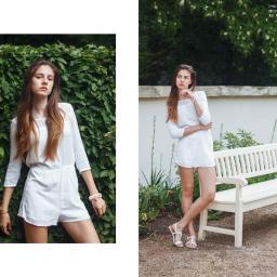 Veludo - Portret młodej kobiety w białym kombinezonie z koronką, pozującej na tle zielonego żywopłotu i białej ławki w parku.