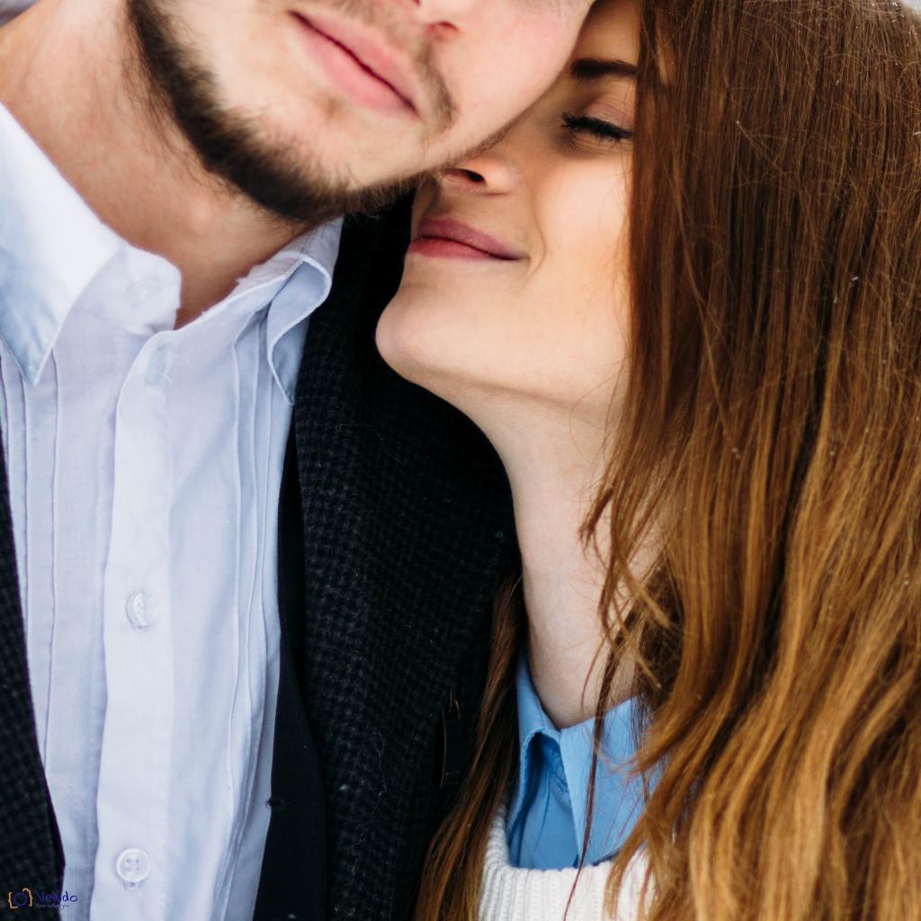 Zbliżenie na profil uśmiechniętej kobiety z zamkniętymi oczami, opierającej się o mężczyznę z brodą, oboje w eleganckich strojach.