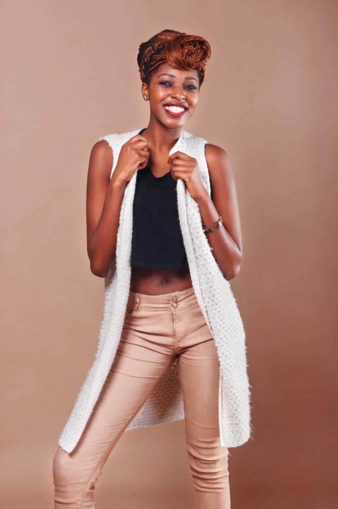 Uśmiechnięta, ciemnoskóra kobieta w białej, dzierganej kamizelce, czarnym topie i beżowych spodniach na jednolitym, beżowym tle.