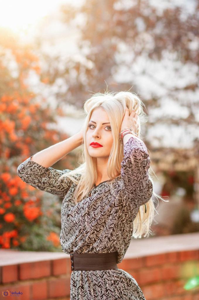 Portret kobiety z blond włosami, w sukience w geometryczne wzory, z czerwonymi ustami, pozującej na tle jesiennej roślinności w słoneczny dzień.