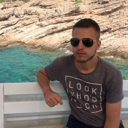 Młody mężczyzna w okularach przeciwsłonecznych, siedzący na ławce z widokiem na skaliste wybrzeże i turkusową wodę.