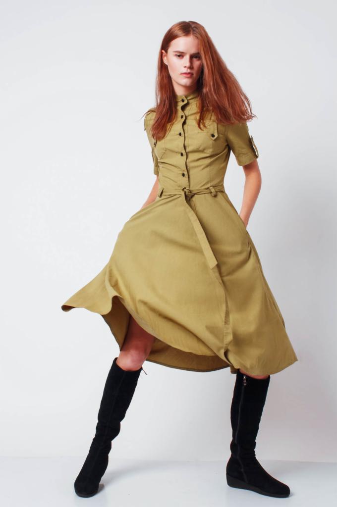 Modelka w ruchu prezentuje khaki sukienkę koszulową z paskiem i czarne kozaki na białym tle.