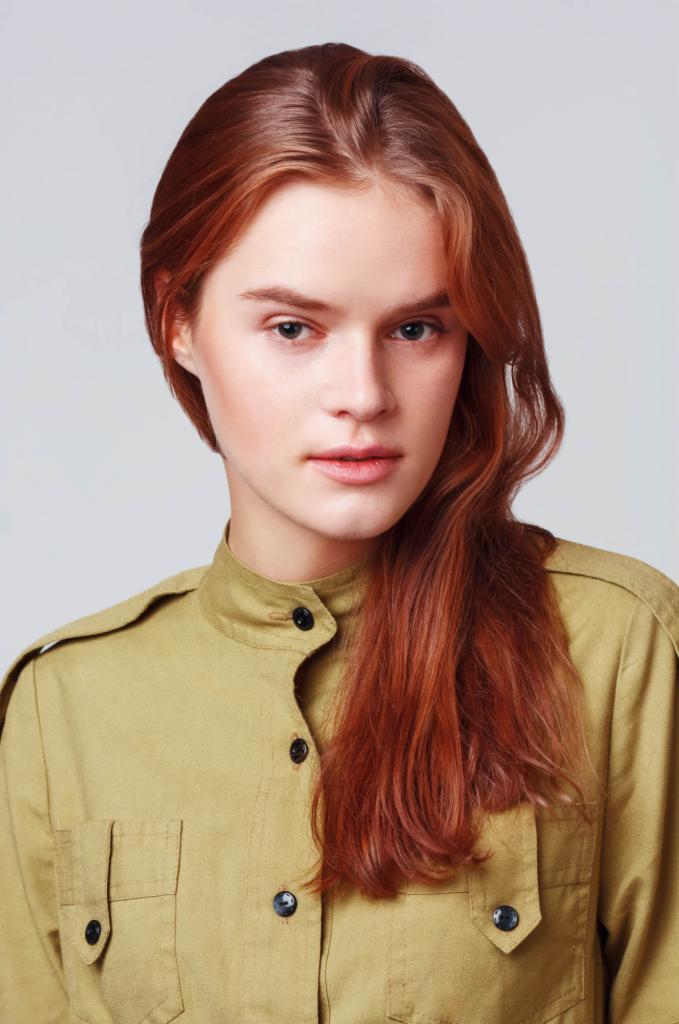 Portret młodej kobiety z rudymi włosami w oliwkowej koszuli z kieszeniami i czarnymi guzikami na neutralnym tle.