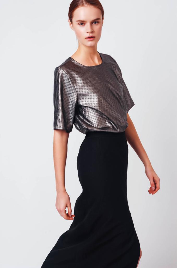 Kobieta pozuje w studyjnym oświetleniu, ubrana w metaliczną bluzkę z krótkim rękawem i czarną, dopasowaną spódnicę maxi z rozcięciem.