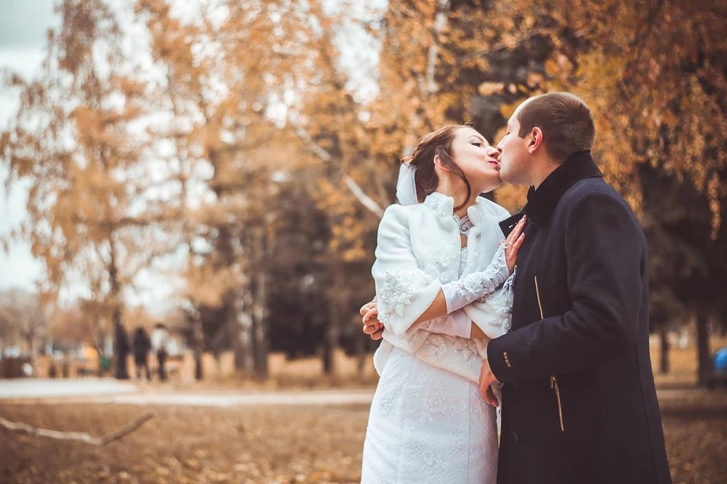 Pocałunek nowożeńców w jesiennej scenerii parku w Warszawie, panna młoda w białej sukni z bolerkiem i koronkowymi rękawiczkami, pan młody w ciemnym płaszczu.