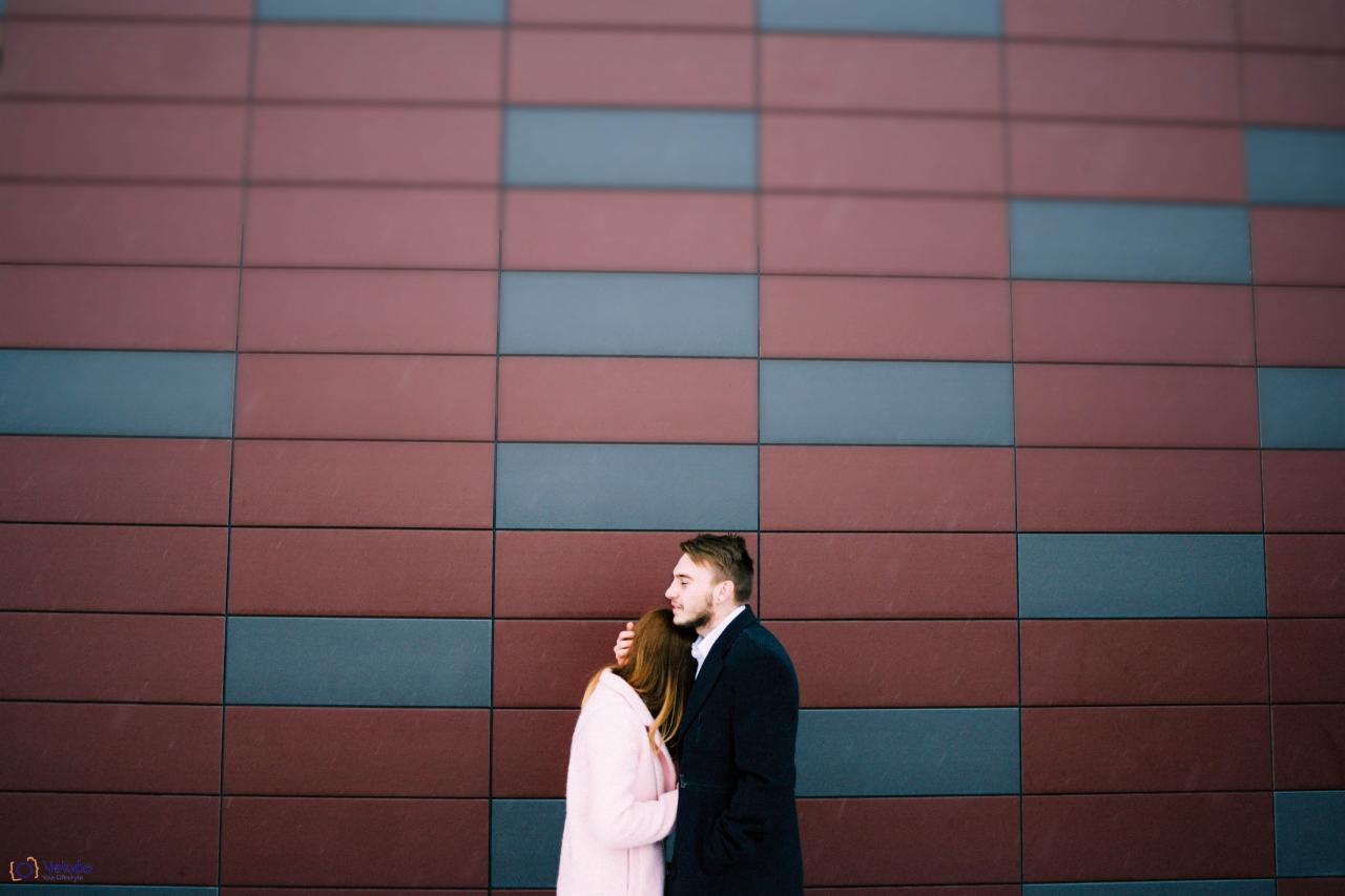 Para obejmuje się na tle ściany z prostokątnych, bordowo-szarych płytek. Mężczyzna w czarnym płaszczu, kobieta w jasnoróżowym płaszczu.