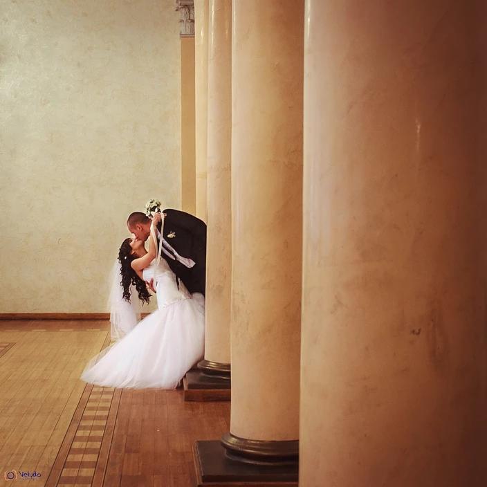 Pocałunek nowożeńców w eleganckim wnętrzu z kolumnami, panna młoda w białej sukni, pan młody w czarnym garniturze, delikatne oświetlenie