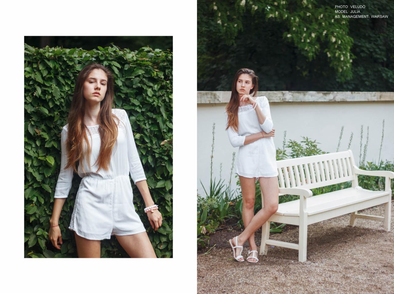 Portret młodej kobiety w białym kombinezonie z koronką, pozującej na tle zielonego żywopłotu i białej ławki w parku.