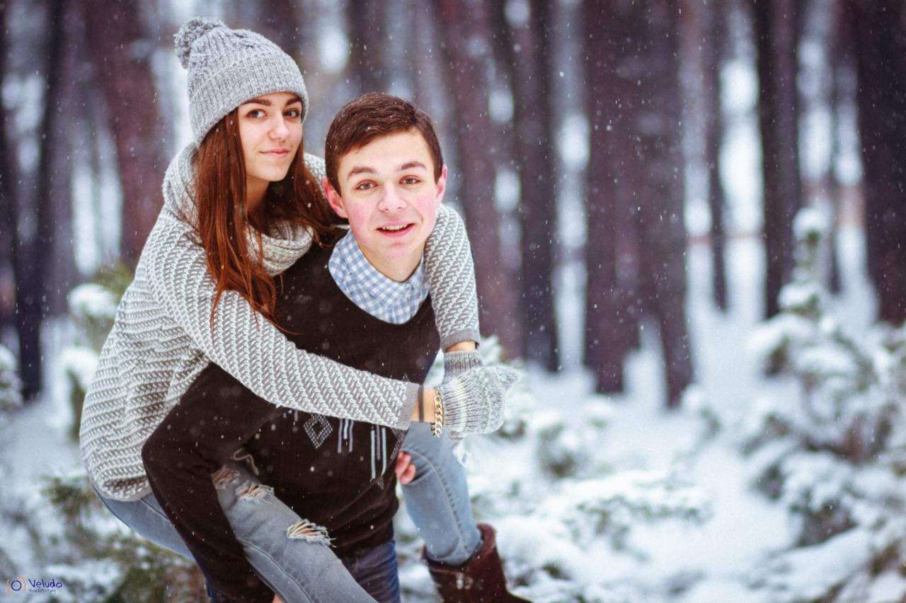 Młoda para w zimowej scenerii: dziewczyna w szarym swetrze i czapce na barana u chłopaka w czarnym swetrze, śnieg pada, las w tle.