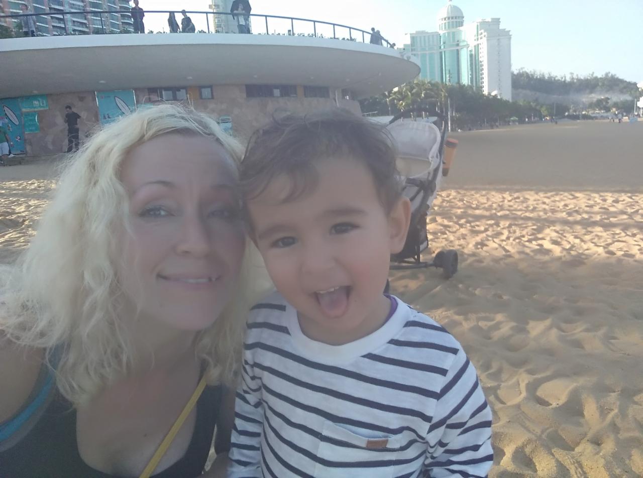 Kobieta z blond włosami i mały chłopiec w paski na plaży z budynkami w tle. Chłopiec wystawia język, a kobieta się uśmiecha. Selfie na tle piasku i wózka.