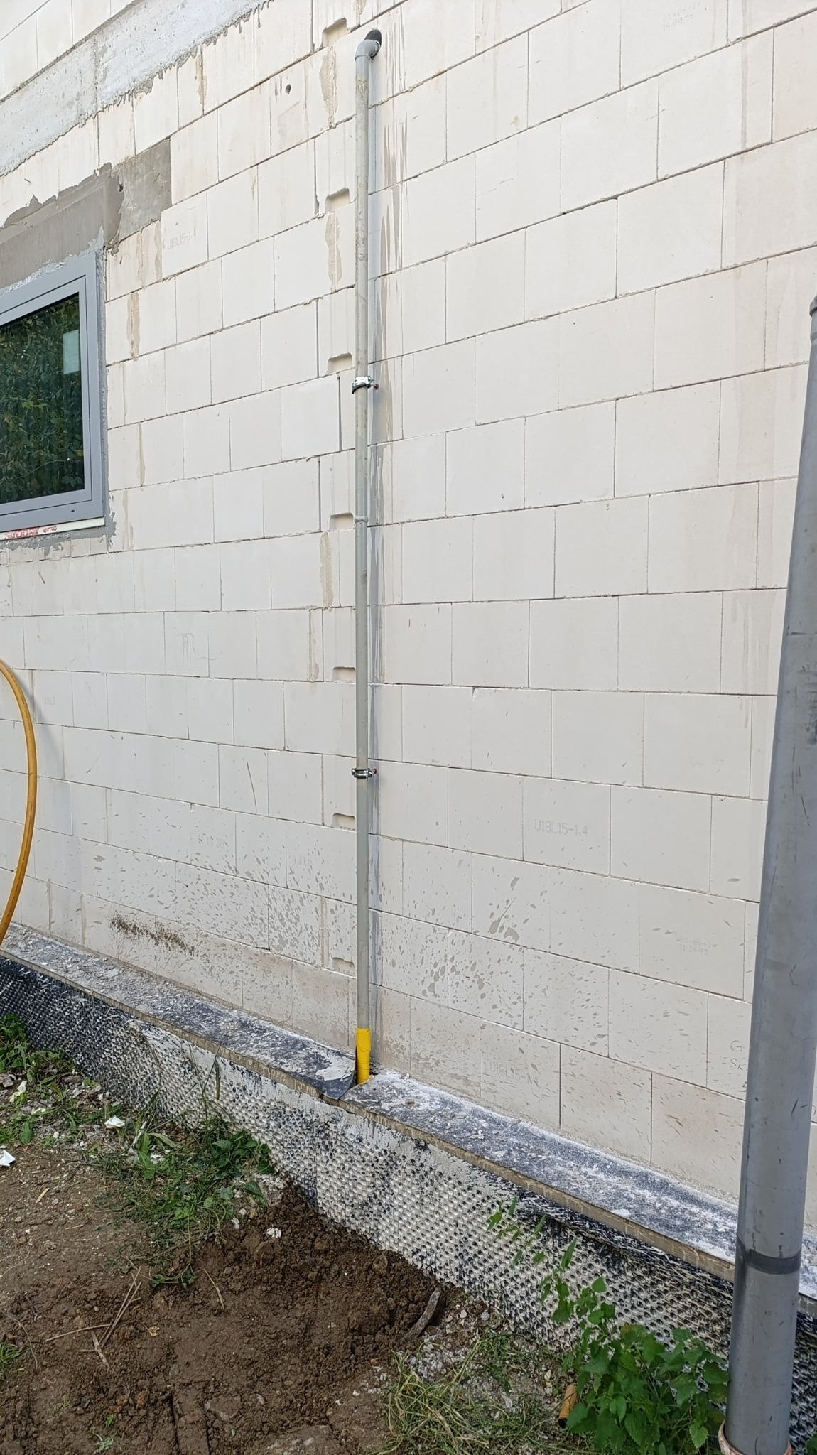 Szara rura pionowa przymocowana do ściany z bloczków betonowych, widoczne ślady po kuciu w ścianie, fragment okna z szarą ramą w lewym górnym rogu, dół ściany zabezpieczony folią kubełkową.