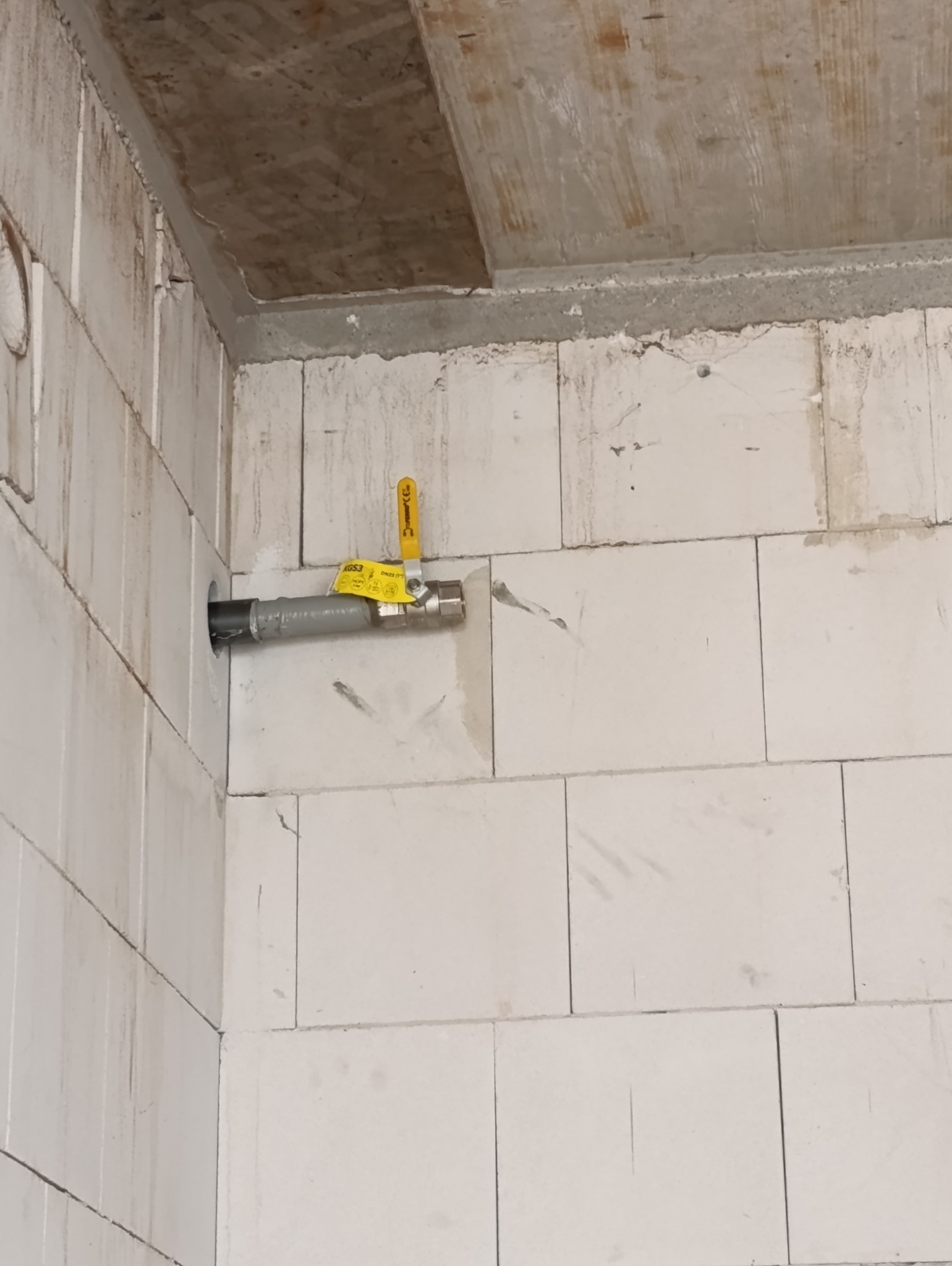 Szara rura gazowa z żółtym zaworem odcinającym wystająca ze ściany z białych bloczków, widoczne ślady po montażu na ścianie i suficie.