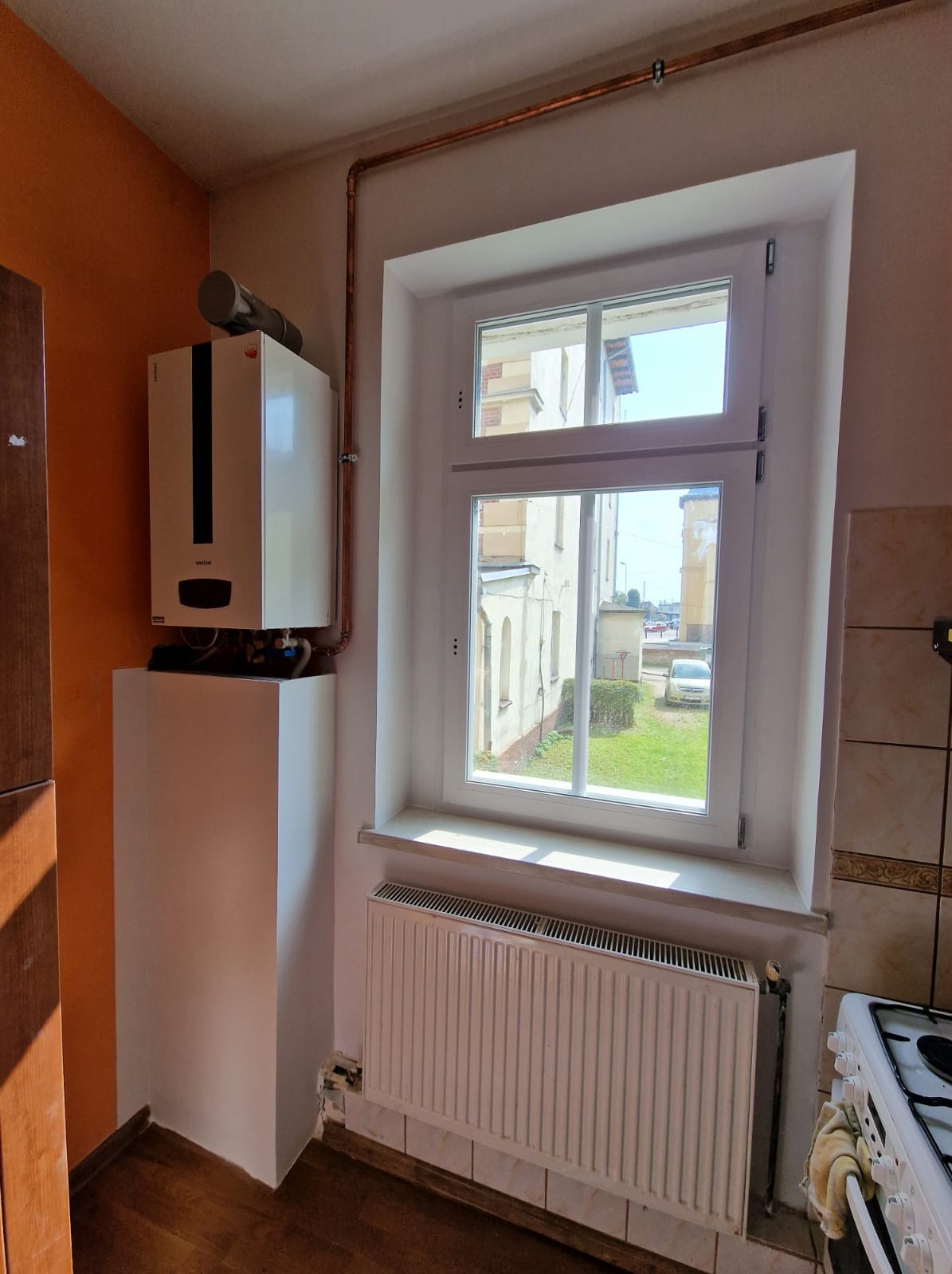 Biały piec gazowy z kominem i szafką u dołu, zamontowany obok okna z widokiem na budynek i trawnik, miedziane rury instalacji grzewczej biegnące wzdłuż ściany.