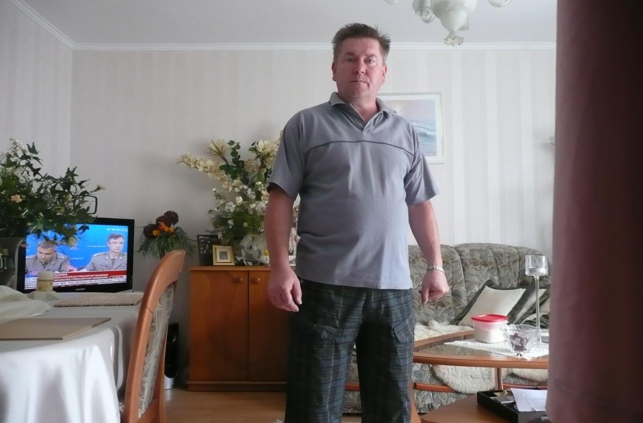 Mężczyzna w szarej koszulce polo i kraciastych spodniach stoi w salonie z telewizorem i kwiatami w tle. Ujęcie od frontu, lekko z dołu.