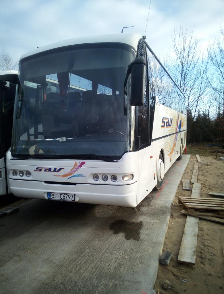 Biały autokar z logo firmy na betonowej platformie, widoczne tablice rejestracyjne i fragment drugiego autobusu po lewej stronie.