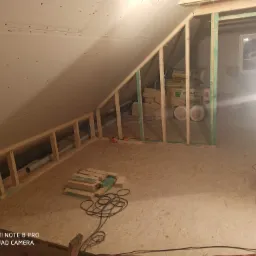 Poddasze w trakcie prac wykończeniowych: widoczna konstrukcja drewniana, ocieplenie w rolkach, płyta OSB na podłodze, częściowo zamontowana płyta karton-gips na skosach, przewody elektryczne...