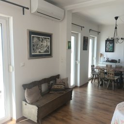 Klimat Instal - Wnętrze jasnego pokoju z zamontowanym klimatyzatorem na ścianie, nad rustykalną ławką z poduszkami; widoczne okna balkonowe, stół z krzesłami i żyrandol w tle.