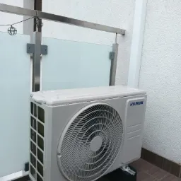 Zewnętrzna jednostka klimatyzacji Hyundai, zamontowana na wspornikach na balkonie z przeszkloną balustradą i dekoracyjnymi lampkami