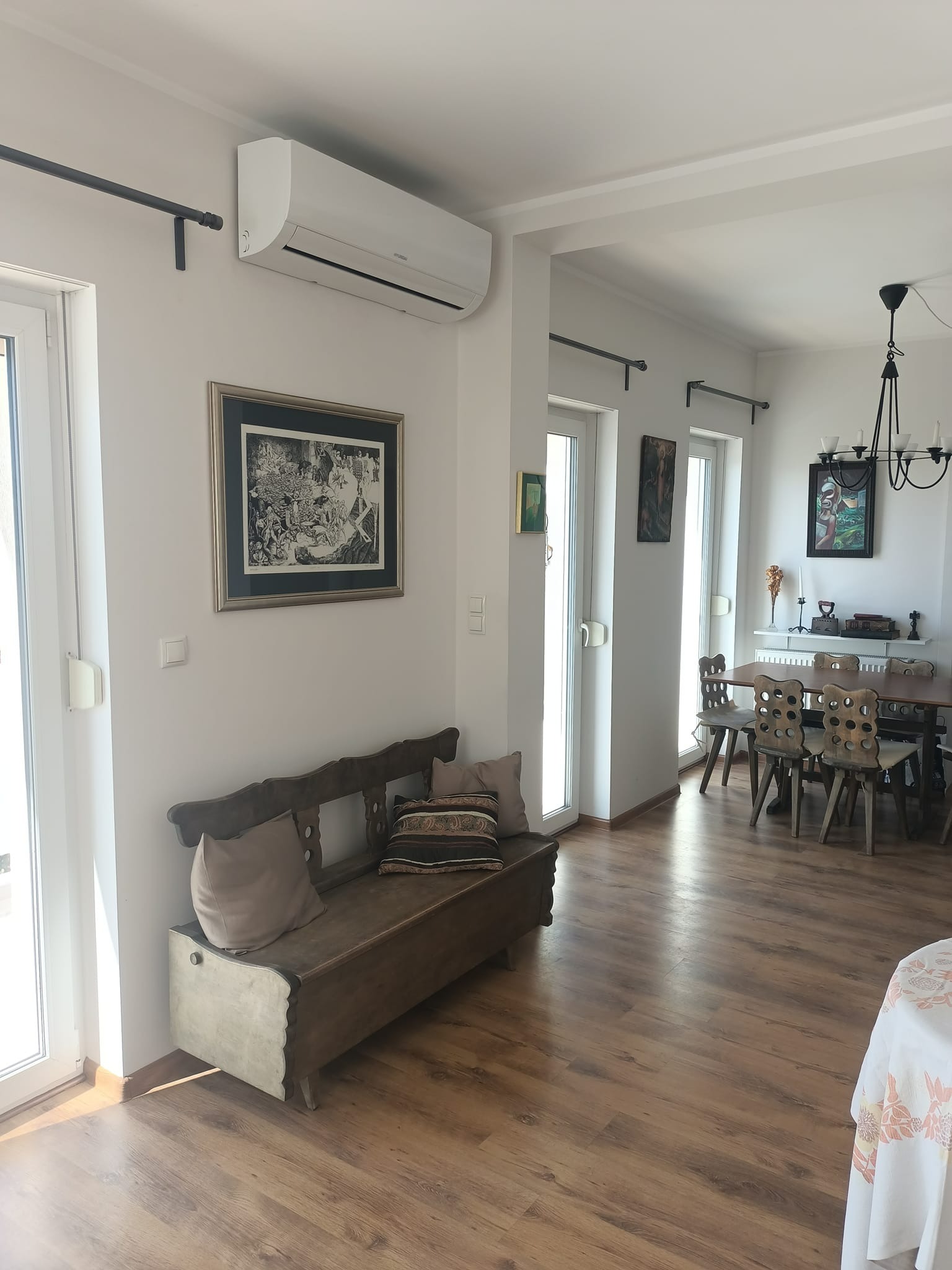 Wnętrze jasnego pokoju z zamontowanym klimatyzatorem na ścianie, nad rustykalną ławką z poduszkami; widoczne okna balkonowe, stół z krzesłami i żyrandol w tle.