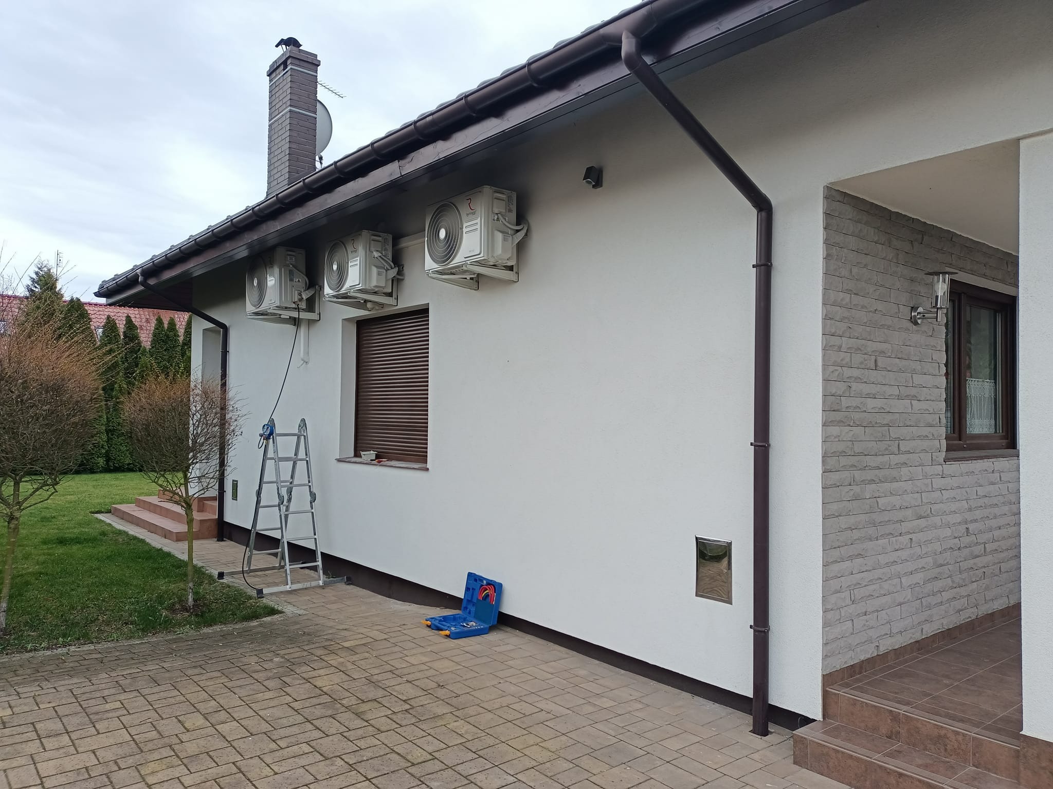 Biały dom z trzema zamontowanymi klimatyzatorami, drabina oparta o ścianę, skrzynka narzędziowa na brukowanym podjeździe.