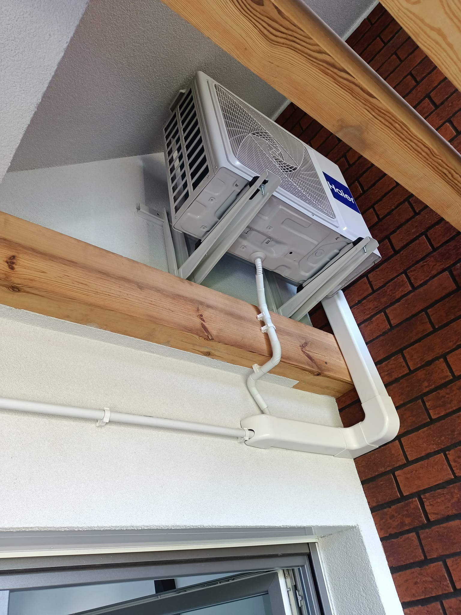 Wewnętrzna jednostka klimatyzacji Haier zamontowana pod sufitem, widoczne rury i kable prowadzone w białych listwach, drewniana belka stropowa i ściana z cegły w tle.
