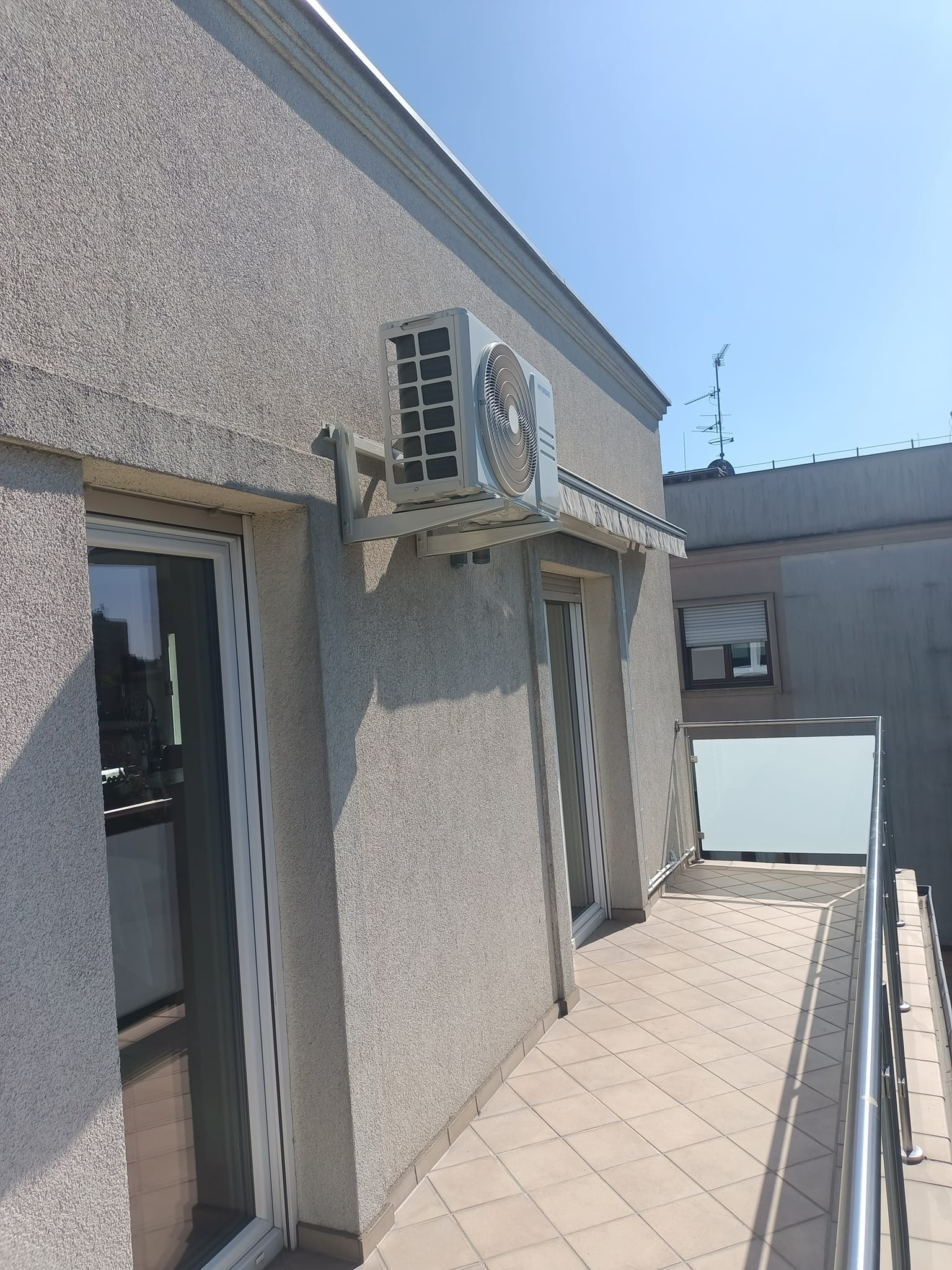 Zewnętrzna jednostka klimatyzacji zamontowana na ścianie budynku z balkonem, widoczna z perspektywy balkonu w słoneczny dzień.