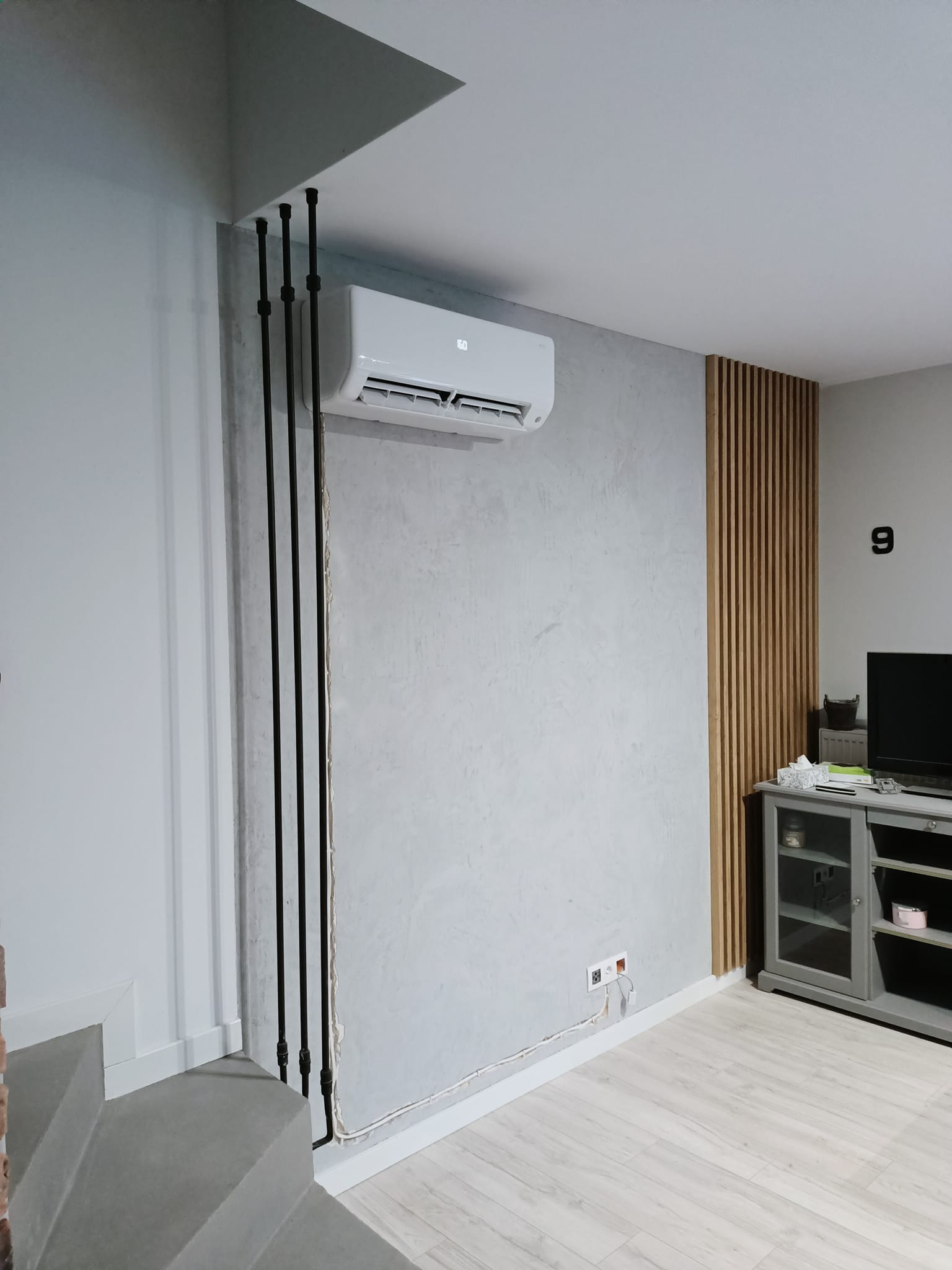 Biały klimatyzator ścienny zamontowany w nowoczesnym salonie z szarą ścianą o fakturze betonu, czarnymi rurami instalacyjnymi i drewnianymi panelami dekoracyjnymi.