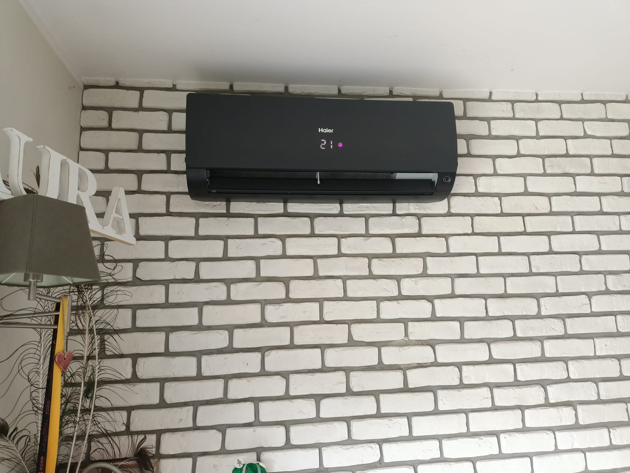 Czarny klimatyzator Haier zamontowany na ścianie z białej cegły, wyświetlający temperaturę 21 stopni Celsjusza.