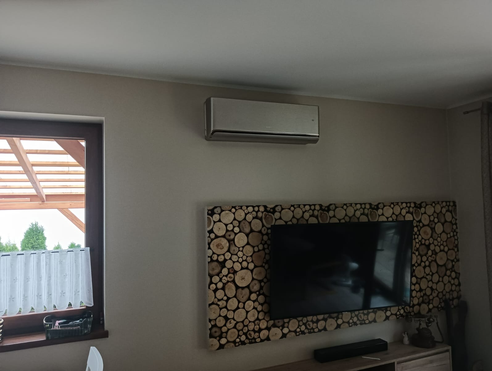 Nowoczesny klimatyzator w kolorze srebrnym zamontowany na ścianie nad telewizorem z drewnianą ramą z przekrojów pni drzew, widok z boku okna.