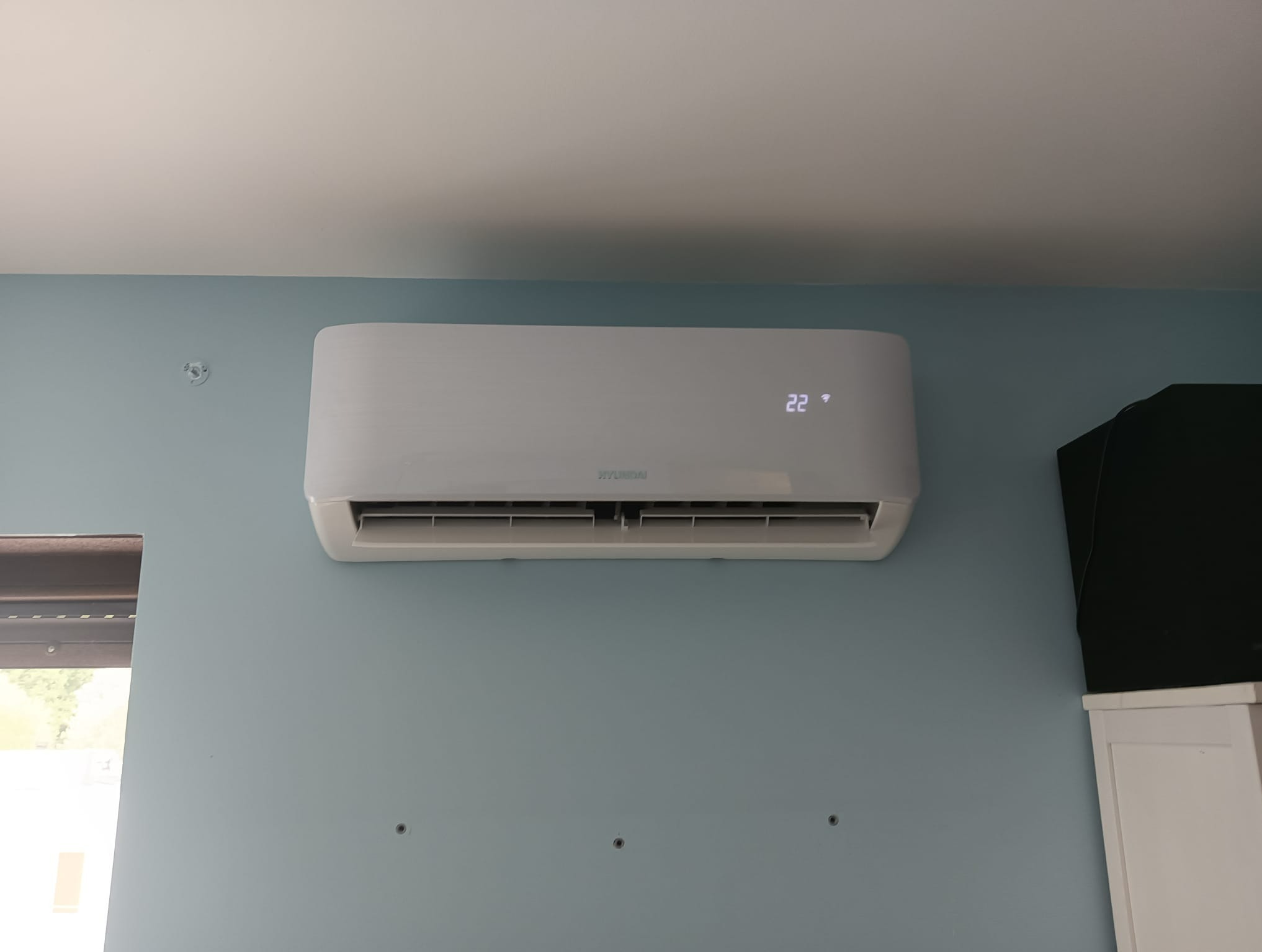 Biały klimatyzator marki Hyundai zamontowany na błękitnej ścianie w pomieszczeniu, z widocznym wyświetlaczem temperatury 22 stopnie.