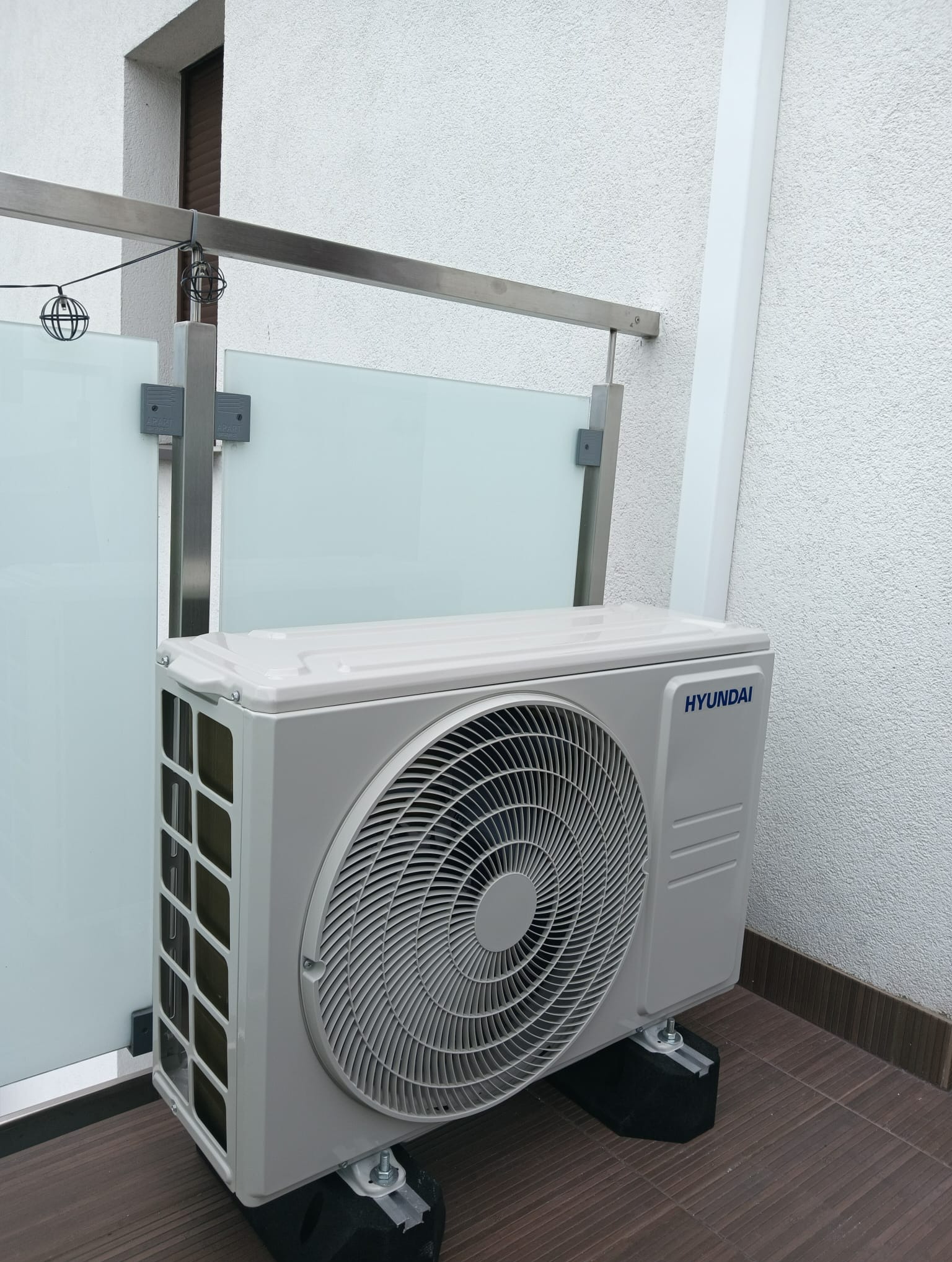 Zewnętrzna jednostka klimatyzacji Hyundai, zamontowana na wspornikach na balkonie z przeszkloną balustradą i dekoracyjnymi lampkami