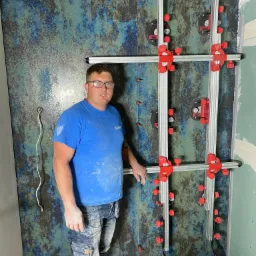 Mężczyzna w niebieskiej koszulce i okularach, ubrudzony zaprawą, stoi na tle ściany z płytek o nieregularnym wzorze w odcieniach niebieskiego i brązu, zamontowanych przy użyciu systemu przyssawek...