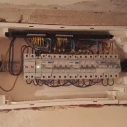 Rozdzielnica elektryczna z zainstalowanymi bezpiecznikami i przewodami w białej obudowie, zamontowana w surowej ścianie z widocznymi śladami tynku.