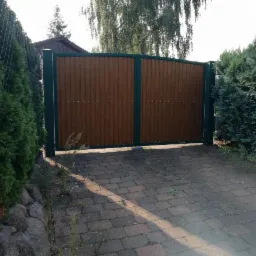 Zamykana brama wjazdowa z paneli drewnopodobnych, osadzona w ciemnozielonej metalowej ramie, z widocznym brukowanym podjazdem i żywopłotem po bokach.