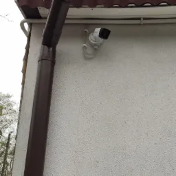Zewnętrzna kamera monitoringu przymocowana do ściany budynku obok brązowej rynny spustowej.
