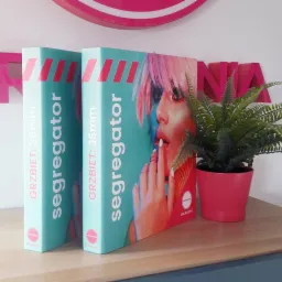 Dwa identyczne segregatory z nadrukiem portretu kobiety w stylu pop-art, ustawione obok doniczki z paprotką na drewnianym blacie.