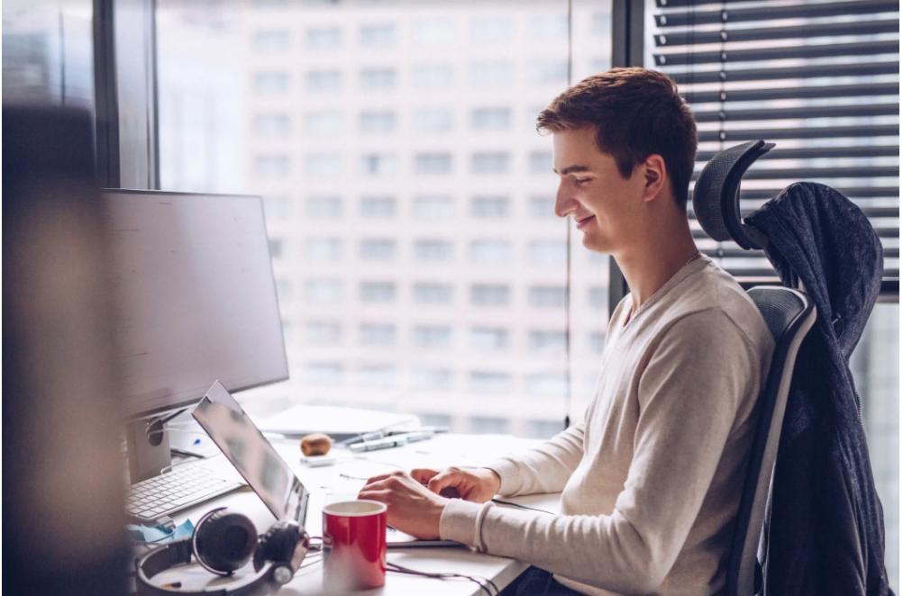 Uśmiechnięty mężczyzna pracuje przy biurku z komputerem i laptopem, z kubkiem i słuchawkami, na tle okna z widokiem na miasto.