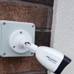 Zewnętrzna kamera monitoringu Hikvision HiWatch Series zamontowana na ceglanej ścianie, widok z boku.