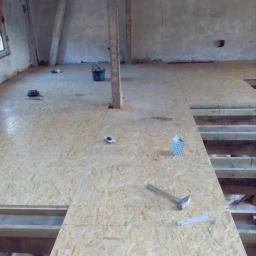 Maksym Popovych - Płyty OSB układane na poddaszu, widoczne narzędzia: młotek, miarka, poziomica oraz wiadro i resztki materiałów budowlanych.