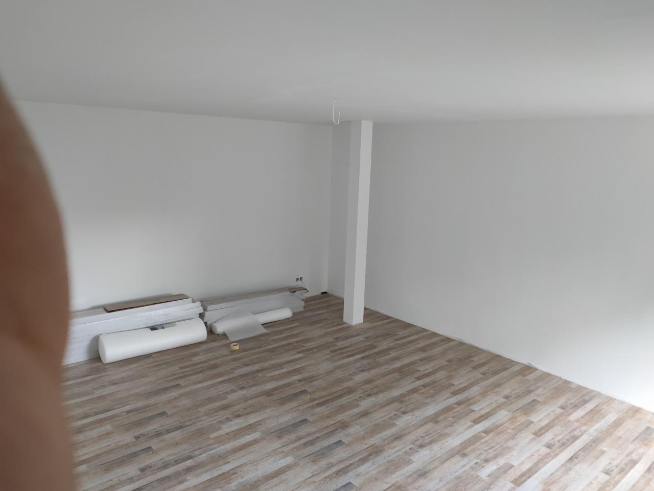 Pusty pokój z drewnopodobną podłogą, białe ściany i kolumna, materiały budowlane w rolkach i paczkach leżące na podłodze przy ścianie, przygotowany do prac wykończeniowych.