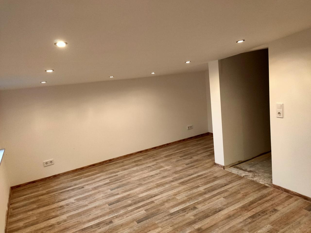 Wnętrze wyremontowanego pokoju z drewnopodobną podłogą, białymi ścianami i sufitowymi lampami punktowymi; widoczne gniazdka elektryczne.