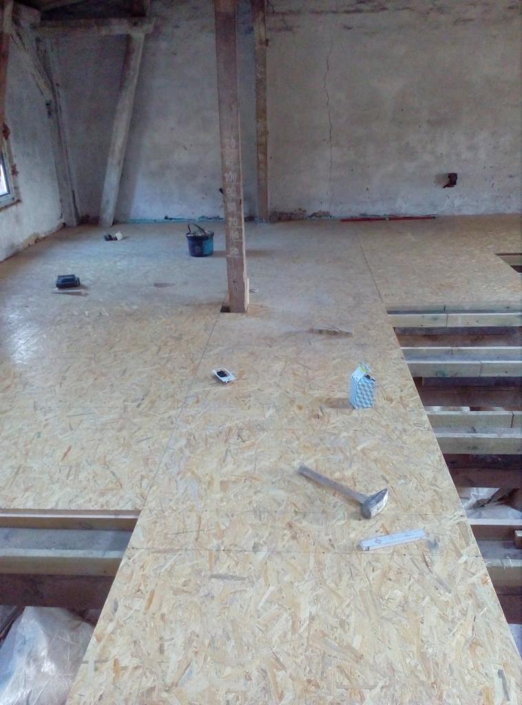 Płyty OSB układane na poddaszu, widoczne narzędzia: młotek, miarka, poziomica oraz wiadro i resztki materiałów budowlanych.